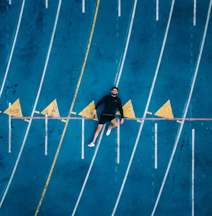 Un homme en tenue de sport est allongé sur une piste de course d'un bleu vif, sillonée de lignes blanches et de marquages triangulaires jaunes. La scène dégage une impression de détente et de contraste.