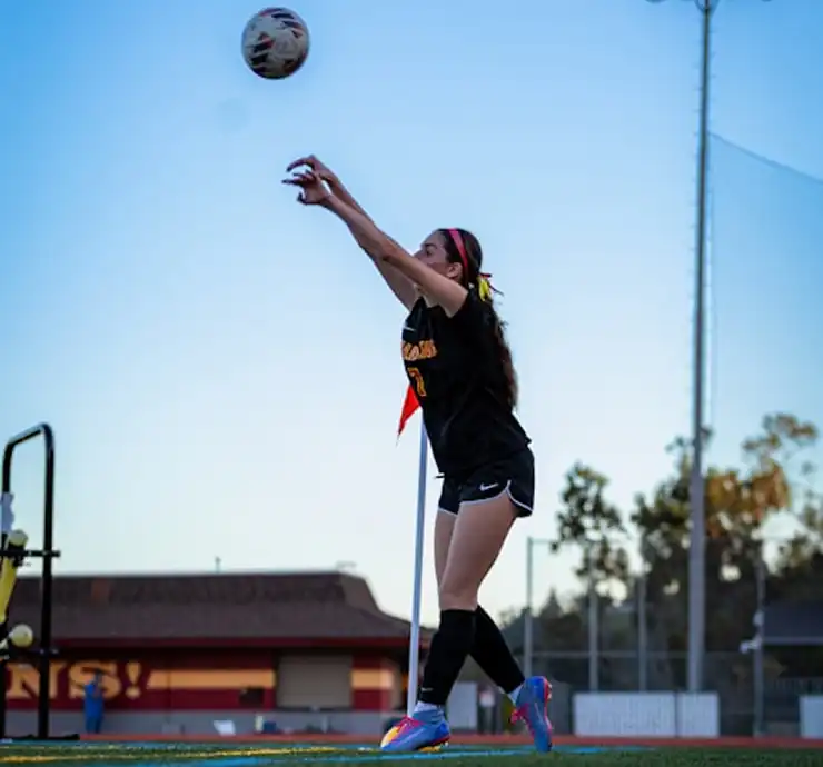 Un footballeur vêtu d'un maillot noir effectue une remise en jeu sur un terrain de football. On le voit en pleine action, le ballon au-dessus de la tête, sur fond de ciel bleu limpide.