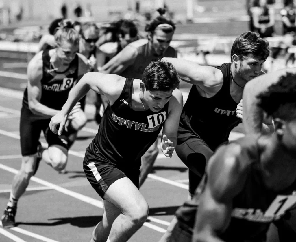 Photo en noir et blanc d'athlètes masculins en train de sprinter sur une piste, faisant preuve d'une concentration intense et d'une énergie compétitive alors qu'ils s'élancent au départ d'une course.
