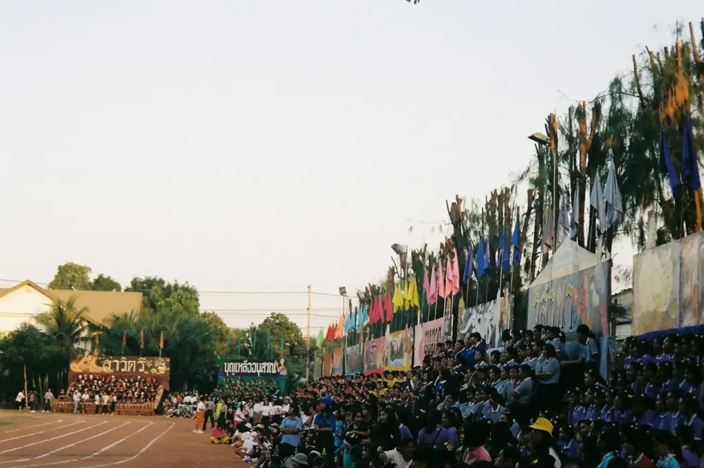 Une foule s'est rassemblée à l'occasion d'un événement sportif ; des drapeaux et des banderoles colorés ornent les tribunes sous un ciel dégagé. L'ambiance est animée et festive.