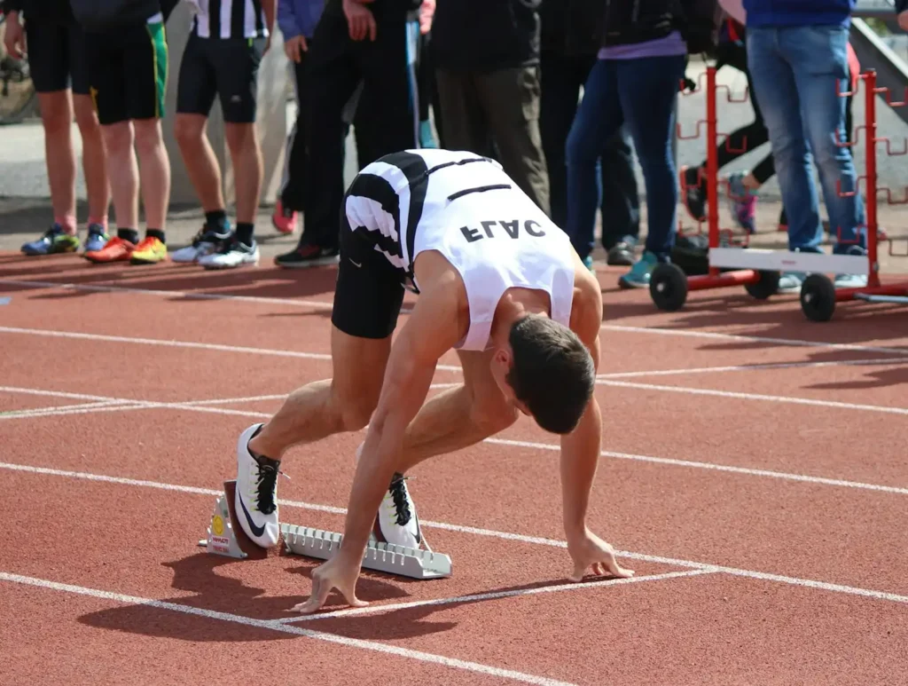 Un athlète en position de départ sur une piste, concentré et tendu, vêtu d'un maillot blanc et noir. Des spectateurs se tiennent à proximité, ajoutant à l'atmosphère de suspense de la scène.