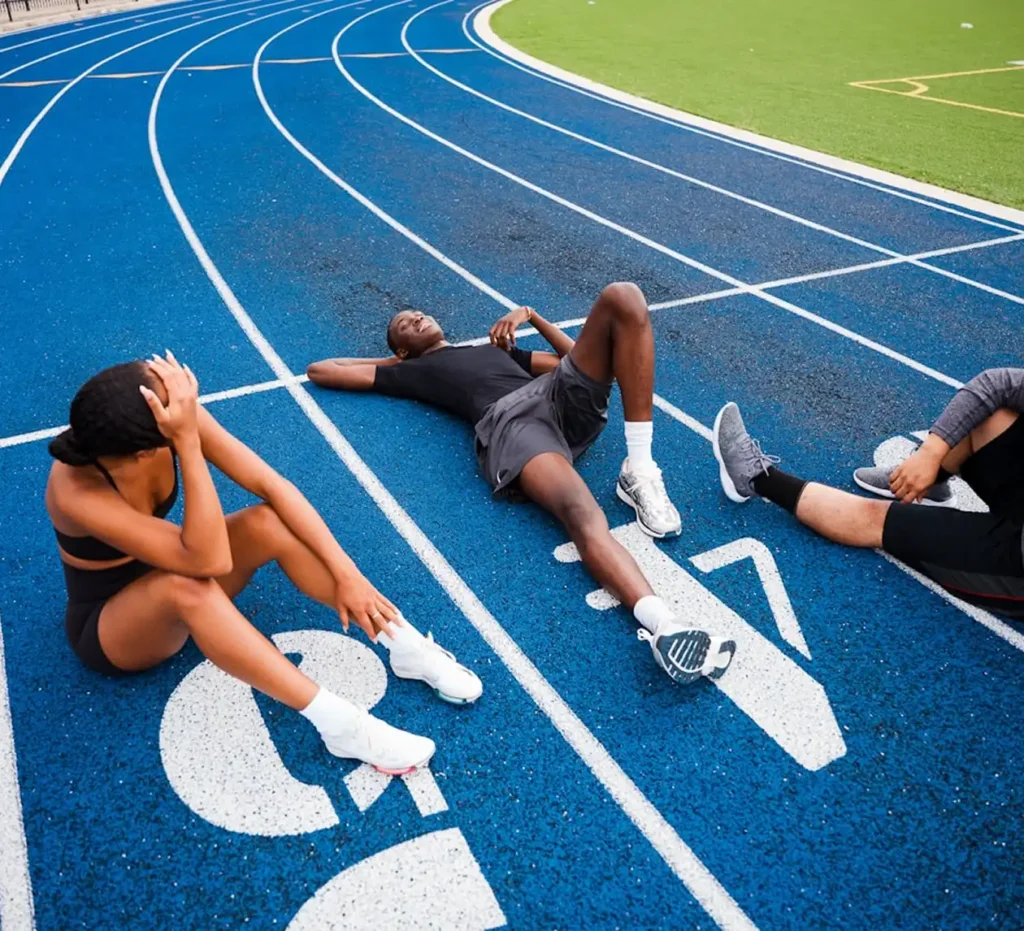 Trois athlètes en tenue de sport se reposent sur une piste de course d'un bleu éclatant. Deux d'entre eux sont assis, la tête baissée, tandis que le troisième est allongé sur le dos, l'air épuisé mais satisfait.