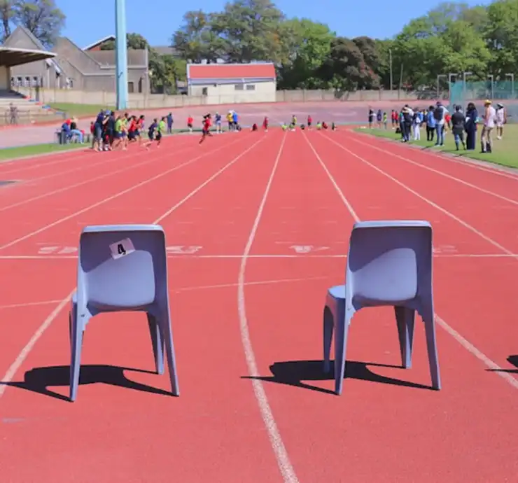 Deux chaises grises placées sur une piste d'athlétisme rouge sont tournées vers une foule lointaine. Sous un ciel bleu limpide, la scène dégage un sentiment d'attente et de sérénité.