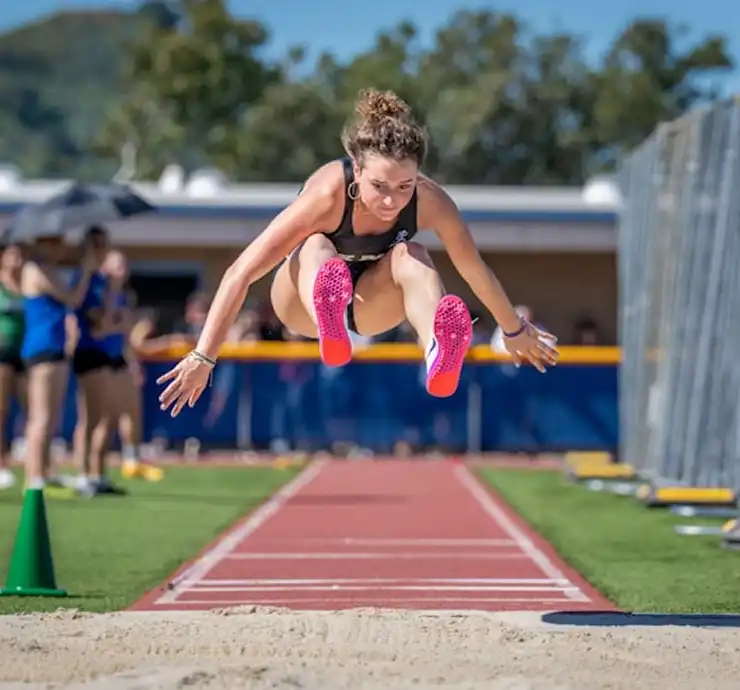 Une athlète en plein saut en longueur, chaussée de baskets rose vif, l'air concentré. Journée ensoleillée, piste et spectateurs en arrière-plan.